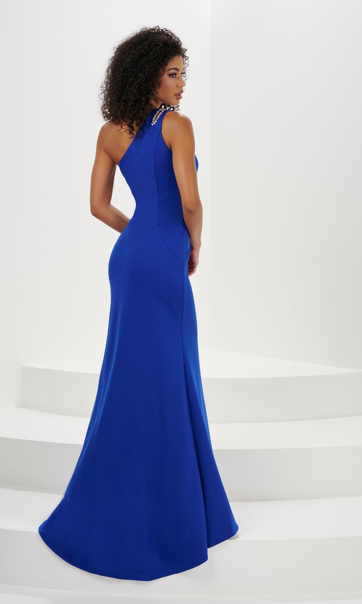 A woman with curly hair wears the Long Prom Dress 14176 by Panoply, a royal blue fitted one-shoulder gown, as she stands on white steps facing away from the camera.